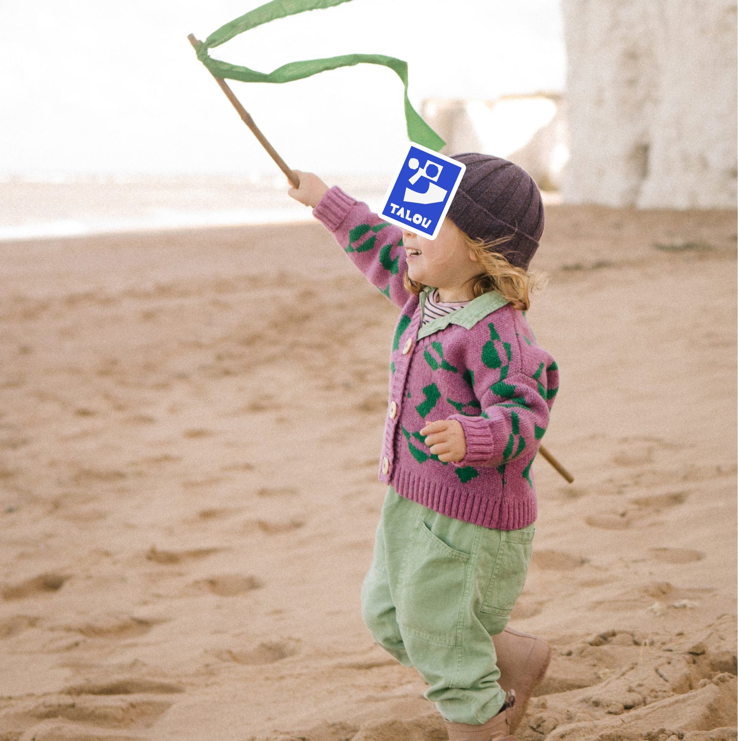 Child running on beach with a flag, wearing a pink and green wool gardigan.