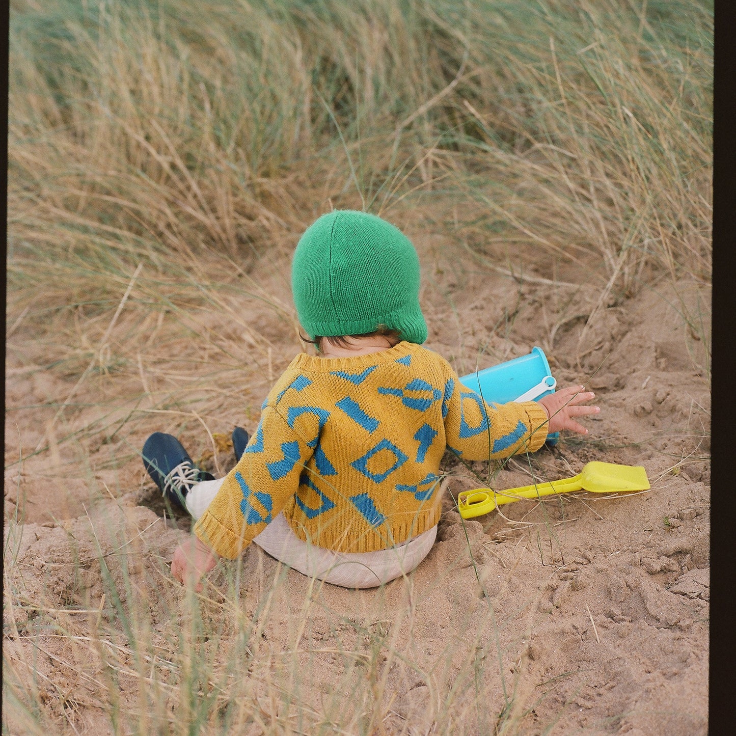 Baby wearing yellow and blue wool cardigan in jacquard with bucket and spade on beach.